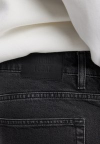 Mustat farkut, joissa on mattamusta nahkaleima, jossa on kohokuvioituna "AMERICA TODAY". Kankaassa on vähän rakennetta ja näkyvät ompeleet.