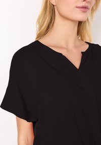 Femme blonde portant un haut noir à manches courtes avec une petite encolure en forme de V, montrée des épaules aux lèvres.