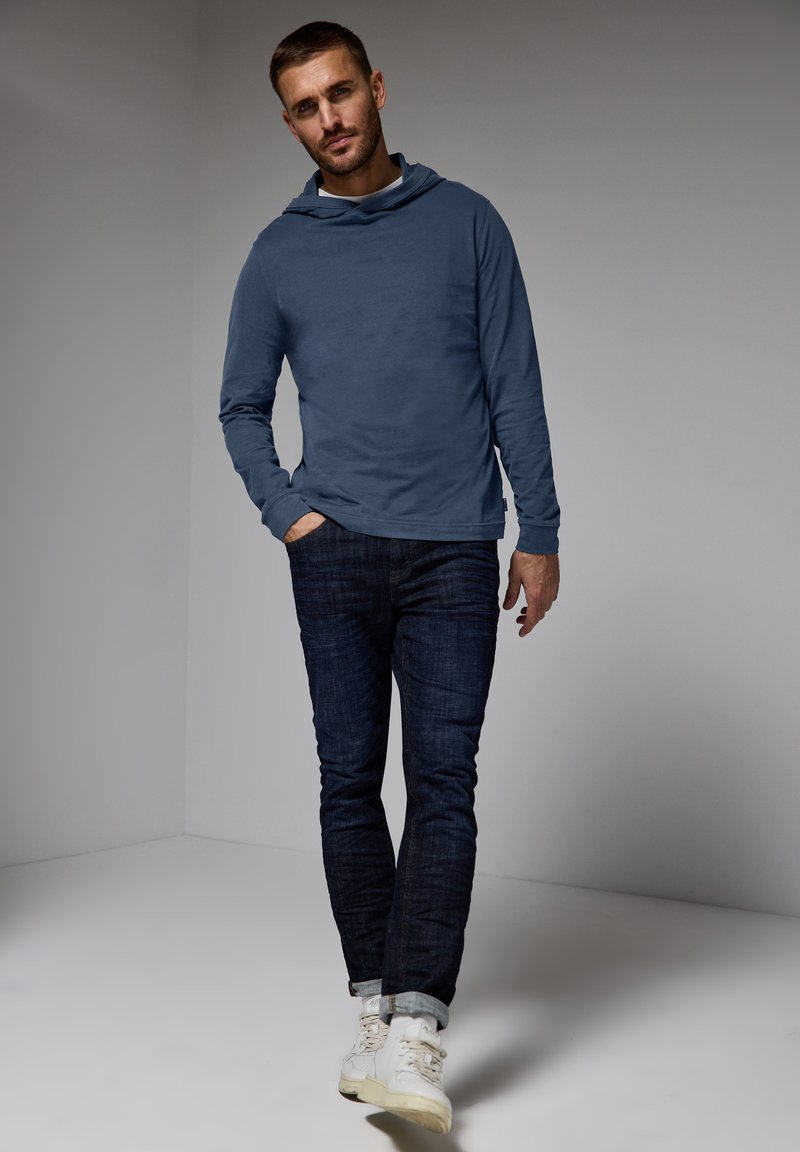 Ljusblå långärmad hoodie, mjuk tyg med krage, kombinerad med mörka denimjeans. Vita sneakers ger kontrast och detaljer.