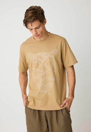 Giovane uomo che guarda verso il basso, indossa una maglietta color cuoio con un disegno astratto bianco di un gufo e pantaloni marroni a quadri, mani nelle tasche.