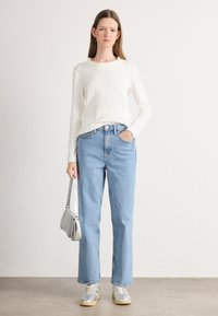 Licht crèmekleurig langemouwen shirt gecombineerd met hoge taille, losse lichtblauwe jeans. Model draagt zilveren sneakers en een kleine zilveren tas.