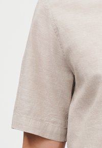 Μπεζ λινό πουκάμισο με κοντά μανίκια στον άνω βραχίονα και τον ώμο ενός ατόμου, που δείχνει την υφή του υφάσματος και τις λεπτομέρειες των ραφών.