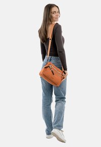 Oranžová, měkk�á látková crossbody taška s dvěma zipovými kapsami a odnímatelným popruhem, kterou drží osoba v džínách a s dlouhým rukávem.