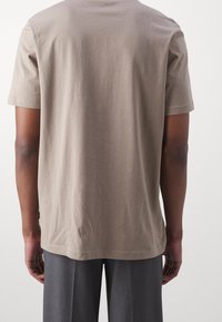 Osoba v jednoduše béžovém tričku s krátkým rukávem a šedých kalhotách zobrazená zezadu na neutrálním pozadí.