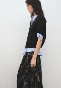 Čierny sveter s výstrihom do V vrstvený cez svetlomodrú pruhovanú košeľu s golierom, v kombinácii s dlhou károvanou sukňou v odtieňoch čiernej a hnedej.