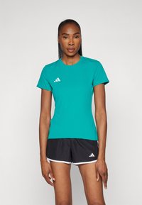 Zöld sportpóló rövid ujjal, kerek nyakkivágással és fehér Adidas logóval a bal mellrészen. Fekete nadrággal párosítva, amelynek fehér szegélye van.
