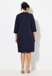 Navyblaues Kleid mit Dreiviertelärmeln, figurbetontem Design und knielanger Schnitt. Glatte Textur und Nahtdetails auf der Rückseite. Getragen mit beigen Absätzen.