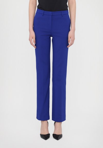 Pantalon bleu ajusté avec une coupe droite, texture lisse et passants de ceinture, associé à des chaussures à talons pointus noires.