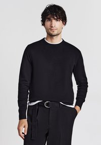 Uomo con capelli ricci scuri che indossa un maglione nero sopra una camicia bianca e pantaloni neri con una cintura di tessuto, in piedi contro uno sfondo chiaro e semplice.