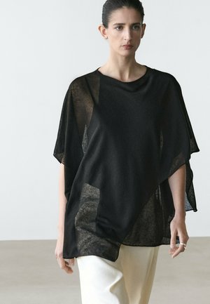 Donna con indosso una maglia nera leggera e trasparente e pantaloni bianchi, in piedi contro uno sfondo chiaro e uniforme con un'espressione neutra.