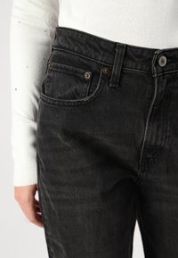Blugi din denim negru cu o textură moale, având două buzunare frontale, accesorii din cupru și un design standard al brâului.