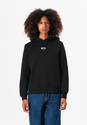 Sudadera con capucha negra, hecha de material suave, con un logo en el frente y puños acanalados. Combinada con jeans de mezclilla azul.
