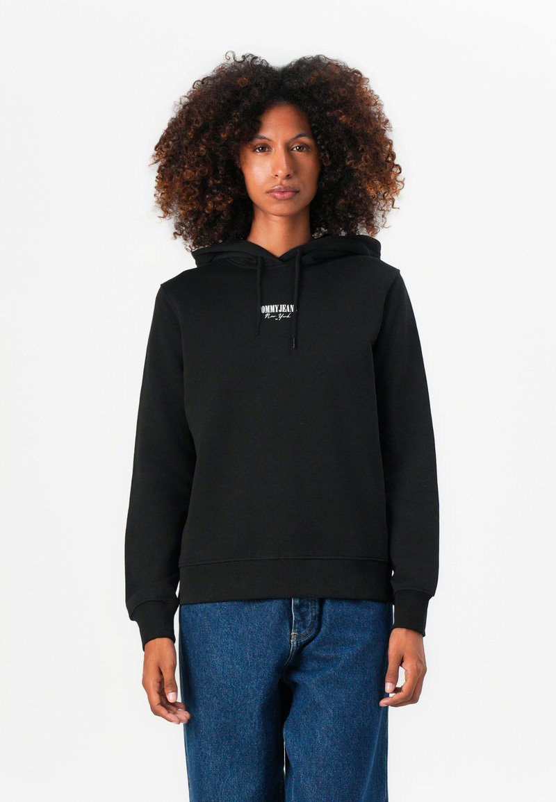 Sweatshirt preto com capuz feito de material macio, apresentando um logotipo na frente e punhos canelados. Combinado com jeans de ganga azuis.