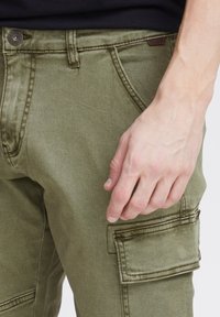 Pantalons cargo vert olive avec taille à bouton, poche latérale et tissu texturé. Une main est visible reposant sur la cuisse des pantalons.