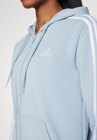 Ljusblå zip-up hoodie i mjukt material, med en framficka, vita ränder på ärmarna och en broderad logotyp på bröstet.