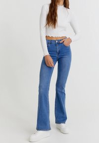 Top blanco de manga larga recortado con dobladillo en forma de ola, combinado con jeans azules acampanados. Tela de mezclilla clara, textura suave y detalle de bolsillo frontal.