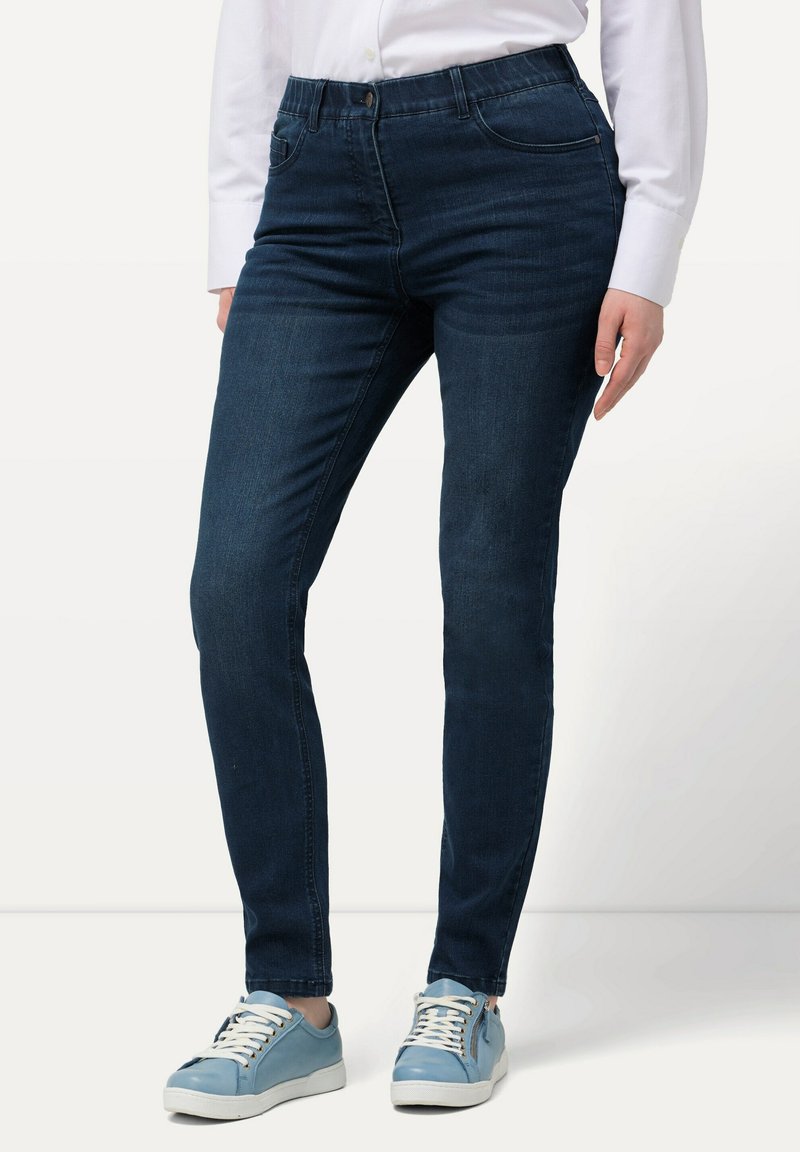 Tmavě modré skinny džíny s hladkou texturou, designem s pěti kapsami a poutky na opasek, nošené s světlomodrými teniskami a bílou košili.