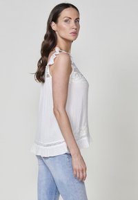 Haut sans manches blanc avec des bretelles ondulées, tissu texturé, détails en dentelle et un ourlet évasé, associé à un jean bleu clair.