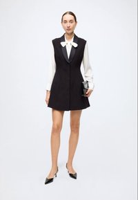 Black sleeveless dress with a satin collar over a white blouse, featuring a fitted design and a flared hem; paired with black heels and a clutch.