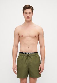 Zöld úszóshort fekete rugalmas derékkal, amelyen a "CALVIN KLEIN" logó látható. A short könnyű és zsinórral zárható.