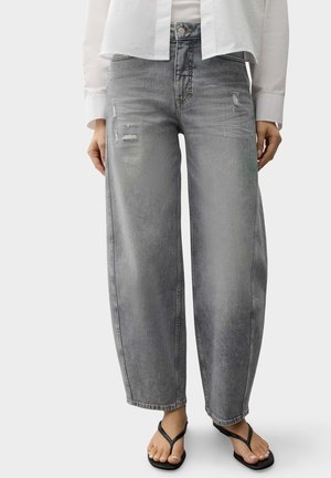 Vrouw draagt lichtgrijze versleten jeans met wijde pijpen, wit overhemd en zwarte slippers, staand tegen een egale achtergrond.