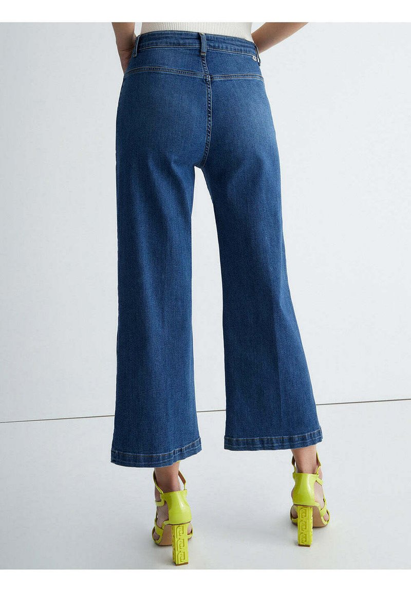 Dżinsowe spodnie o szerokich nogawkach w ciemnoniebieskim kolorze z wysokim stanem. Posiadają czysty wykończenie i wykończone nogawki, w zestawieniu z neonowymi zielonymi sandałami na wysokim obcasie.