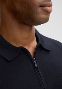 Marineblauwe polo shirt met ritssluiting, gemaakt van een textuurstof en met een kraag. Het shirt heeft een ritssluiting aan de voorkant en een minimalistisch ontwerp.