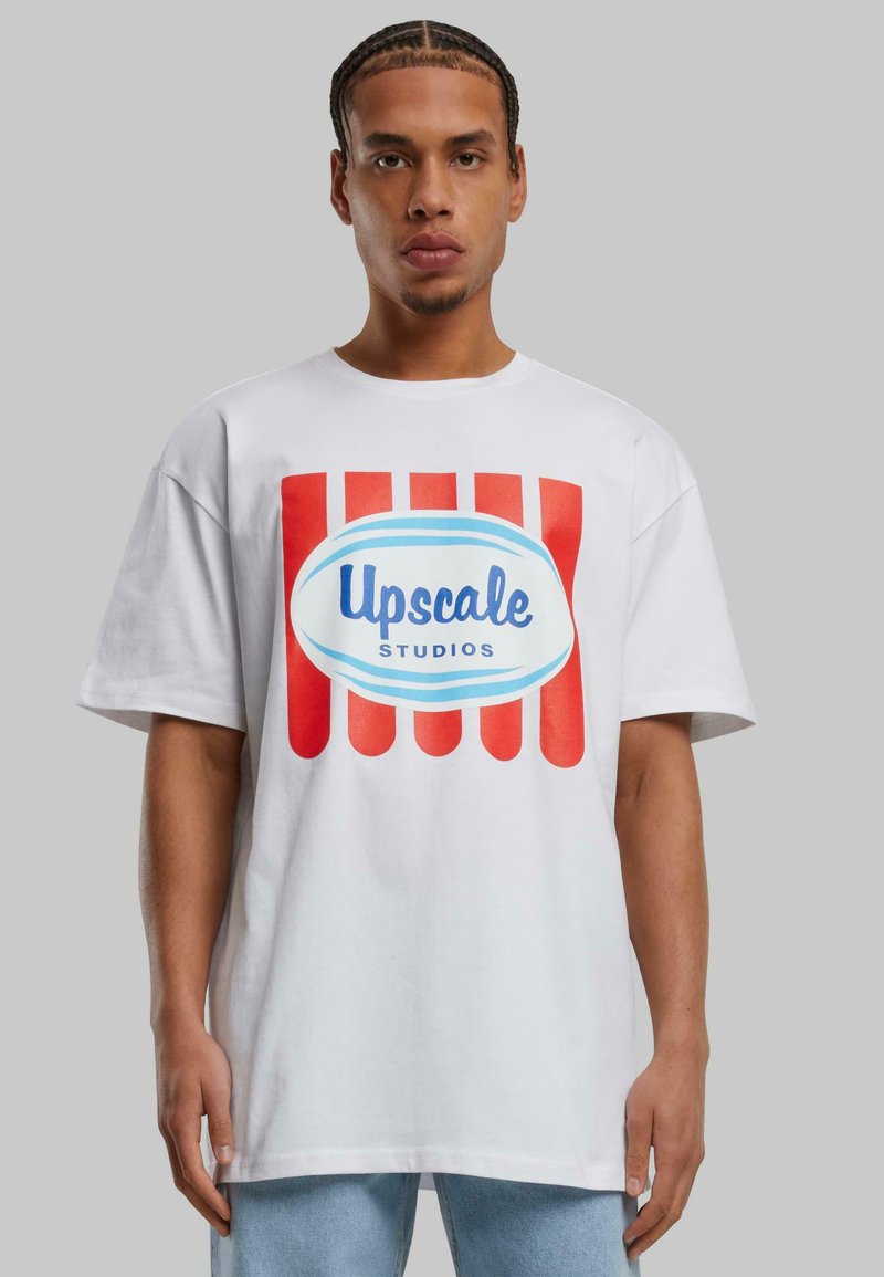 White cotton t-shirt featuring a large graphic in red and blue, reading "Upscale Studios" on a rounded background. Short sleeves, relaxed fit.