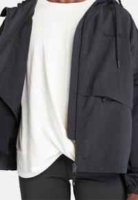 Giacca leggera nera con chiusura a zip, cappuccio e fodera in rete, indossata sopra una maglia bianca. Presenta una tasca laterale e polsini elastici.