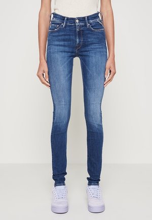 Jeansy niebieskie slim-fit noszone z białymi trampkami na stojącej osobie na jednolitym tle.