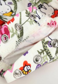 Tissu blanc présentant des illustrations florales et de dessins animés colorés, avec des boutons-pression argentés le long du bord, et une couture légèrement effilochée.