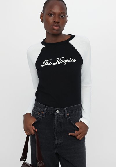 Black and white raglan long-sleeve shirt featuring "The Kyrples" text in white, paired with dark denim high-waisted pants and a burgundy handbag.