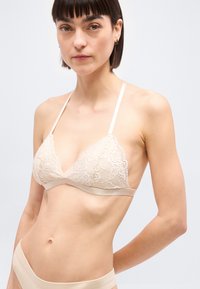 Γυναίκα που φοράει μπεζ δαντελένιο σουτιέν με λεπτές τιράντες και αντιστοιχία σλιπ ψηλόμεσο, σε απλό φωτεινό φόντο.