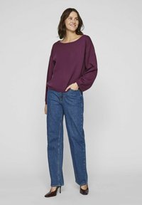 Un pull violet ample avec un décolleté large, associé à un jean bleu à taille haute et coupe droite, et des talons hauts marron.