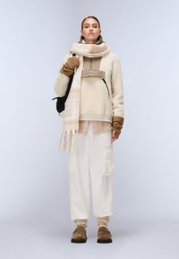 Μπεζ φλις πουλόβερ με τσέπη με φερμουάρ, κρεμ κασκόλ με ρίγες, φαρδιά κρεμ cargo παντελόνια και καφέ παντόφλες με λουράκια.