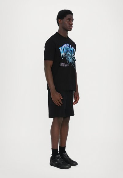 Black cotton T-shirt with blue graphic print, paired with black shorts and black footwear. Model stands on a plain background in profile view.