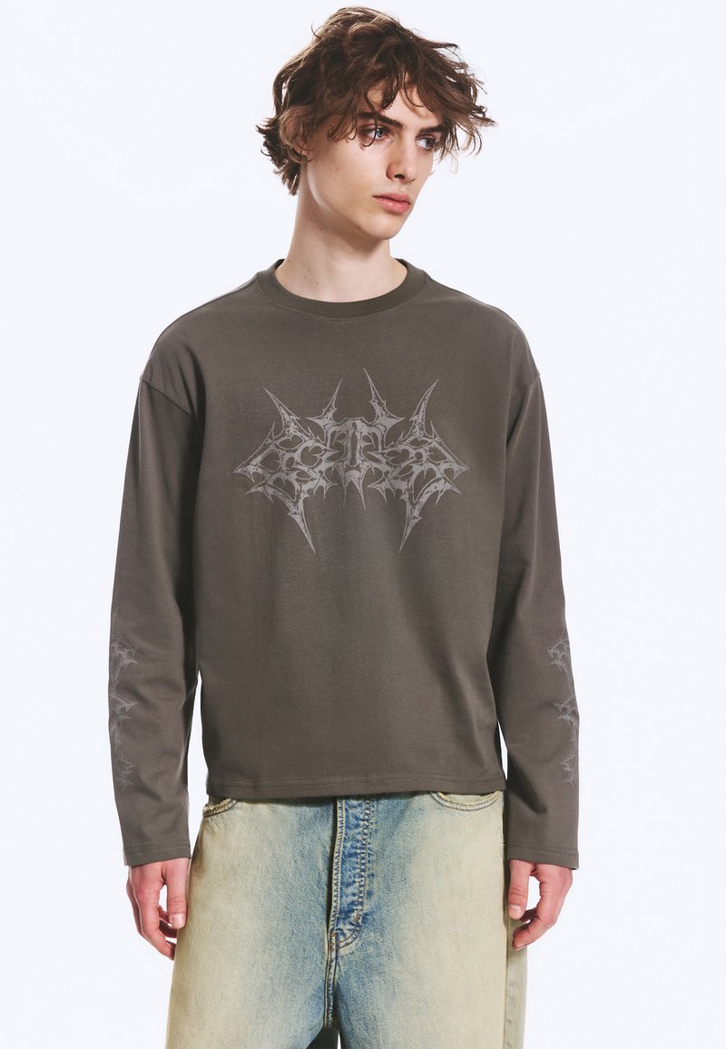 Jeune homme aux cheveux bouclés porte un t-shirt à manches longues gris foncé avec un motif graphique abstrait et un jean bleu clair délavé sur fond blanc.