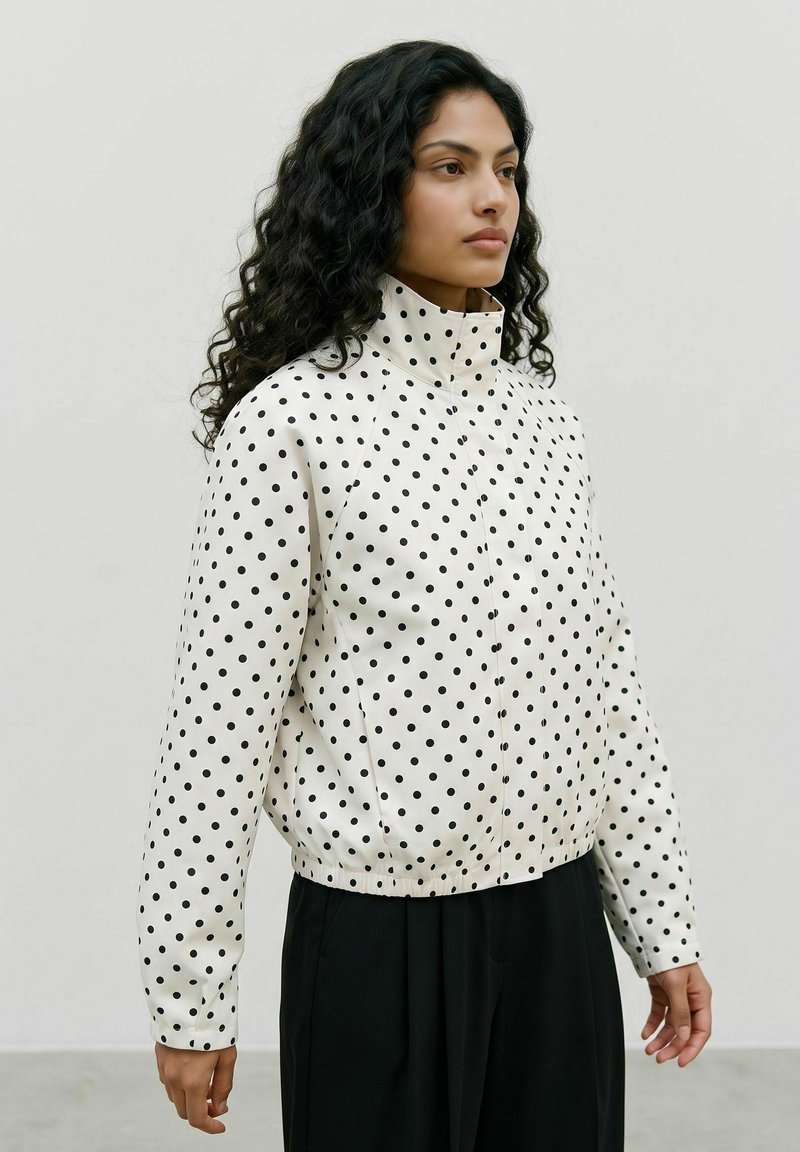 Femme aux longs cheveux bouclés portant une veste blanche à pois noirs et col montant, associée à un pantalon noir ample.