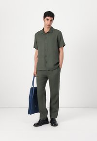 Camicia verde a maniche corte e pantaloni abbinati con una vestibilità su misura. Indossato con scarpe nere e mentre tiene una borsa a tracolla in denim blu.