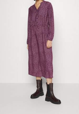 Frau trägt ein langärmeliges violettes Midikleid mit kleinen weißen Punkten und schwarze klobige Lederstiefeletten.