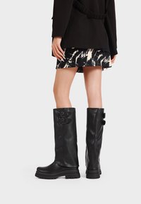 Botas de cuero negro hasta la rodilla con una textura mate, que cuentan con dobles hebillas en el costado y una suela de goma gruesa para mayor tracción.