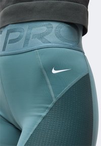 Κοντινή λήψη από πετρόλ κολάν Nike Pro με διάτρητες πλευρικές πάνελ και λευκό λογότυπο Nike στη γοφή.