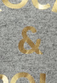 Tessuto grigio con una finitura testurizzata, caratterizzato da grandi lettere dorate metalliche e un simbolo di esperanto. Il design enfatizza una tipografia audace.