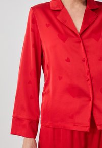 Top in raso rosso con colletto, maniche lunghe e motivi a cuore. Presenta una chiusura con bottoni e una texture liscia.