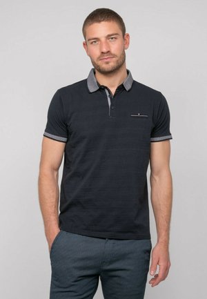 Homme aux cheveux courts portant un polo noir avec un col et des bords de manches à motifs, debout avec une main dans la poche.