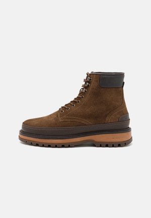 Brown suede ankle boots featuring a thick rubber sole, leather accents, and metal eyelets for laces. Stack heel design for added height.