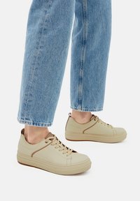 Bežid laced sneakers, mille tekstuuriga pealispind, ümmargune varvas ja pruunid aktsendid. Kombineeritud helesiniste sirgete jalgade teksadega.