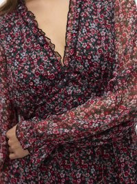 Γυναίκα που φορά διαφανές, μακρυμάνικο φόρεμα με λουλούδια σε κόκκινο, ροζ και λευκό χρώμα, με βολάν στο ντεκολτέ και τις μανσέτες, με τα χέρια σταυρωμένα πάνω στο σώμα.