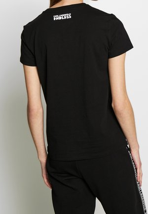 Osoba nosząca czarną koszulkę z krótkim rękawem z napisem „KARL LAGERFELD ENDLESS” na górnej części pleców oraz dopasowane czarne spodnie z paskiem z logo.