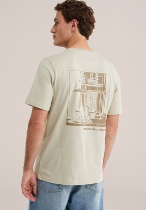 Homme portant un t-shirt beige avec une illustration sépia d’une terrasse de café et le texte « Terraza en la esquina » au dos, associé à un jean bleu.